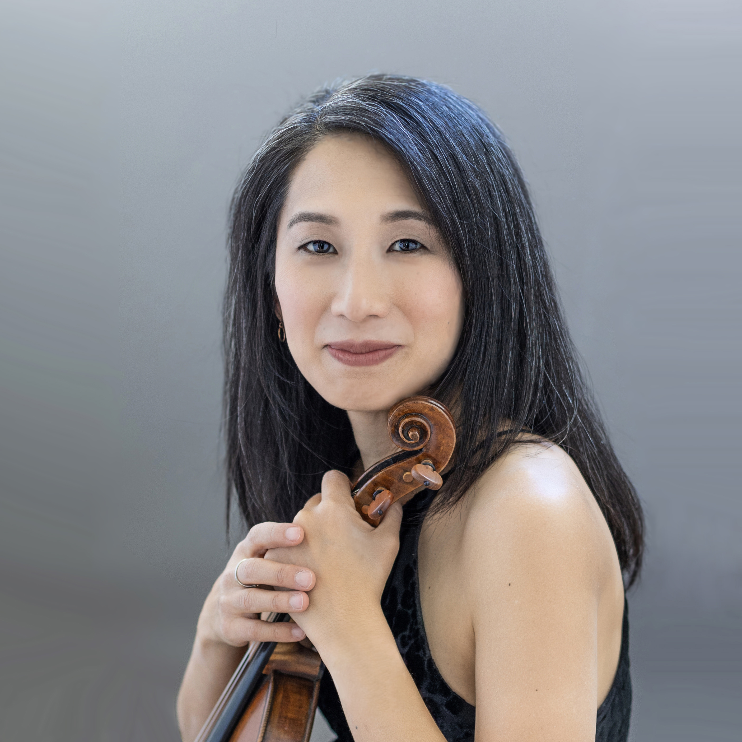 Sarah Kim sits with her violin in front of a gray backdrop facing the camera.